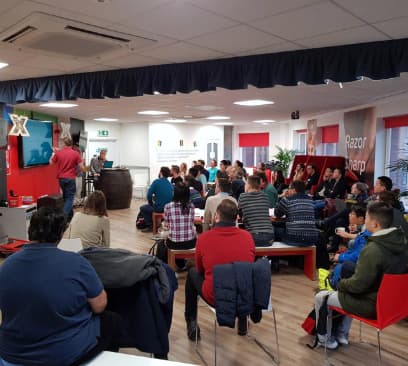 A room full of people all sitting on different seating arrangements listening to two men who are in front of a screen.